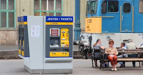 Utazó emberek egy pályaudvaron, tájékoztató táblákkal és utazási információkkal a háttérben.